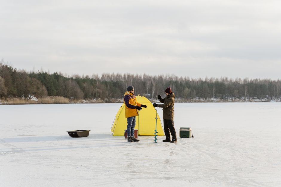 Two anglers ice fishing on a frozen lake with yellow tent and gear in winter.